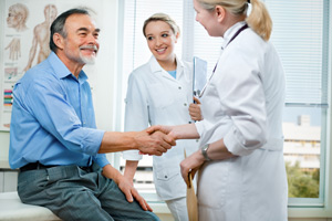 Picture of a male sitting on a doctors table shaking hands with a female Physician as a female nurse is standing there beside them