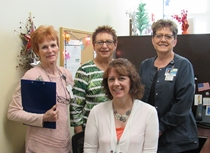 Picture of four females smiling. 
One on the far left side is a older lady with red hair and a pink shirt holding a blue clipboard next to her in the back is a lady with brown hair and glasses with a green striped shirt and the lady to the far right has short brown hair with a blue shirt on, there is a lady in the front with short brown hair and a white shirt with a orange under shirt on. They are in a office type setting