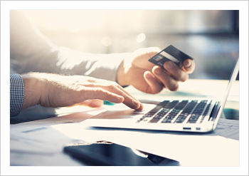 Picture of a man sitting down at a desk with an open laptop, while his hand is on the keyboard and the other hand is holding his debit or credit card. 