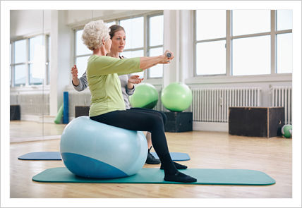 Picture of a Physical Therapist helping a elderly women that is doing workouts while sitting on a yoga ball