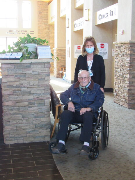 Picture of a femåle Volunteer standing behind a male, elderly patient in a wheelchair in the main Lobby of the hospital.