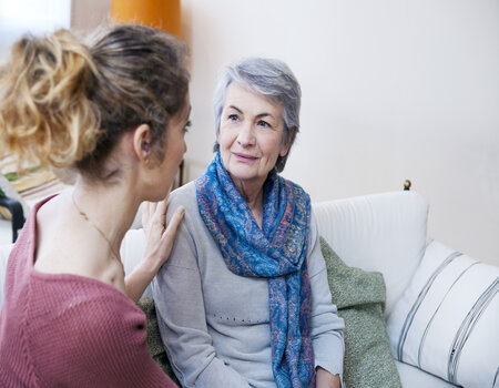 Picture of a female putting her hand on an elderly ladies shoulder. They are both smiling at each other.