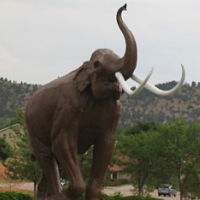 Picture of an statue elephant blowing his trunk. There is woods and hills in the background.